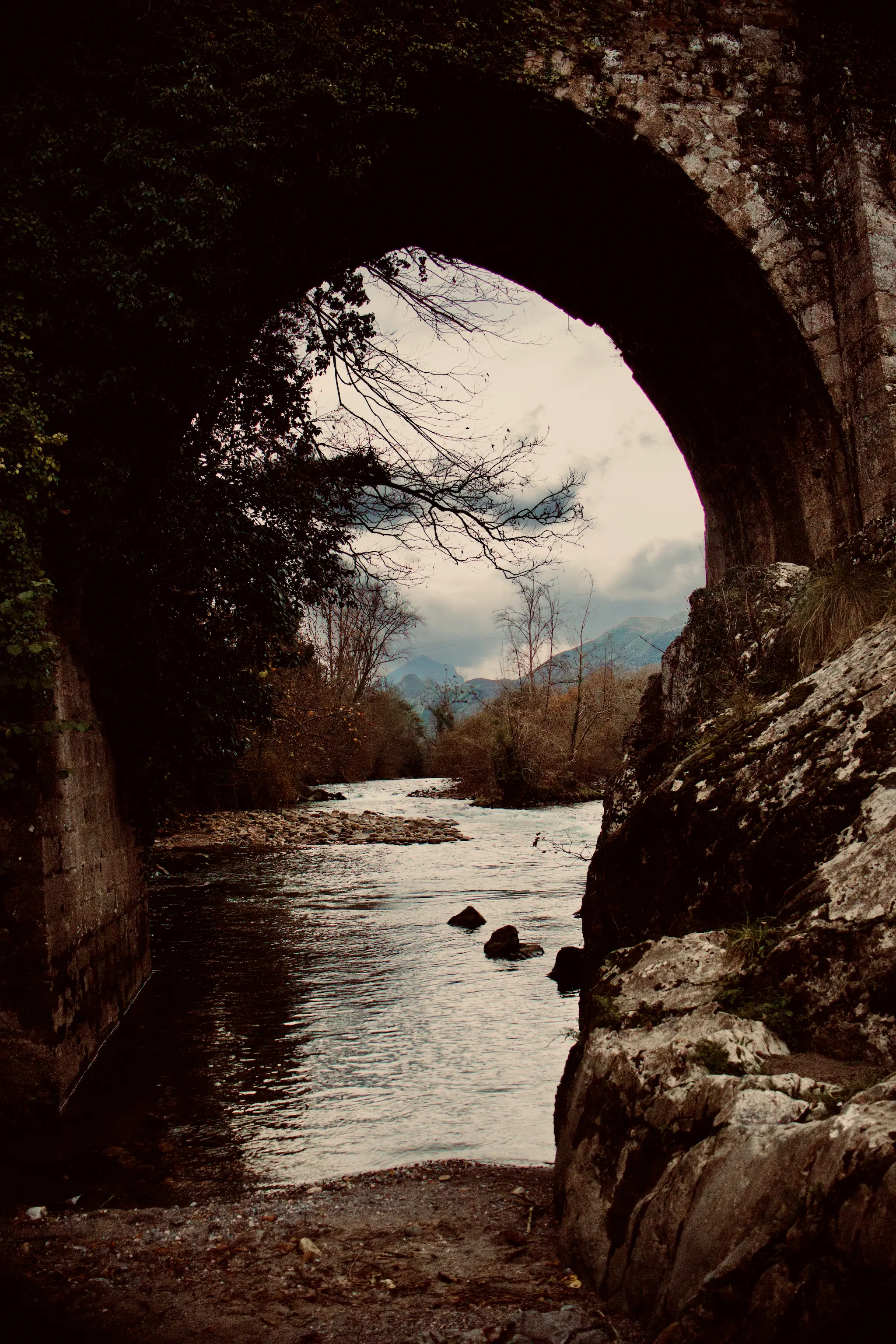 Río de montaña pasando bajo un arco de piedra con árboles desnudos y montañas al fondo en Cangas de Onís Asturias