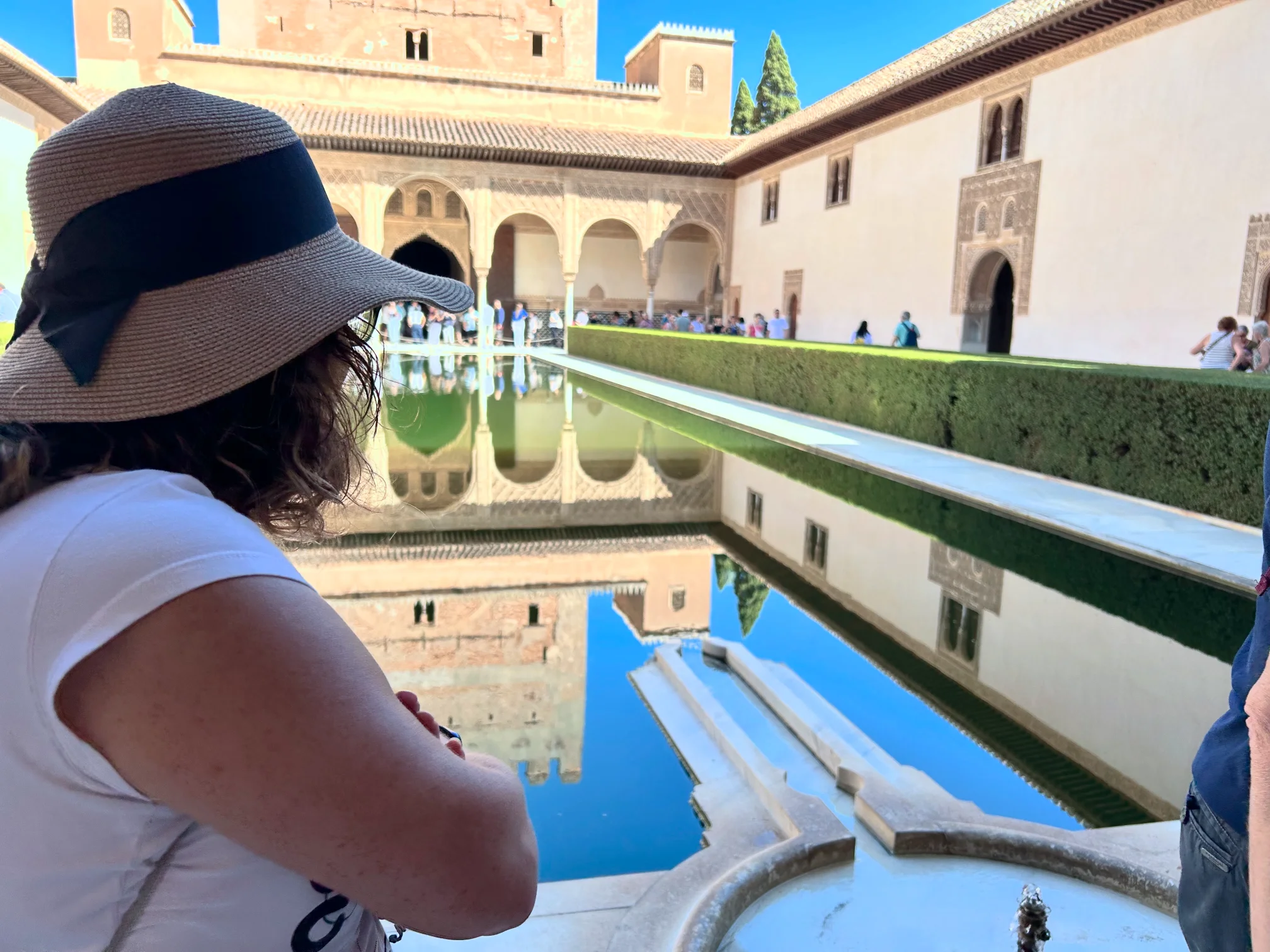 Mi Mujer (Olga) con sombrero contemplando los reflejos del Patio de los Arrayanes en la Alhambra de Granada, con la alberca central en calma y la arquitectura nazarí duplicada en el agua.